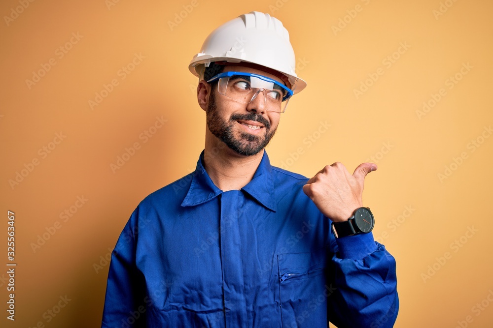 Mechanic man with beard wearing blue uniform and safety glasses over yellow background smiling with happy face looking and pointing to the side with thumb up.