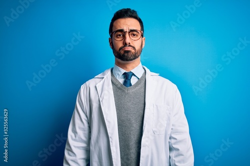 Young handsome doctor man with beard wearing coat and glasses over blue background depressed and worry for distress, crying angry and afraid. Sad expression.