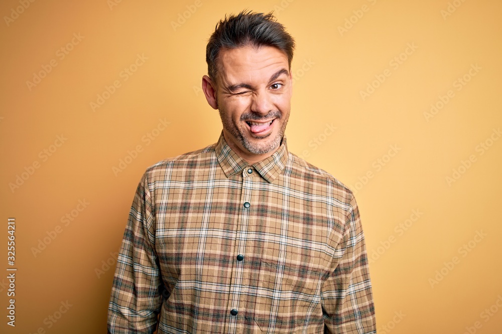 Young handsome man wearing casual shirt standing over isolated yellow background winking looking at the camera with sexy expression, cheerful and happy face. Young handsome man wearing casual shirt standing over isolated yellow background winking looking at the camera with sexy expression, cheerful and happy face.