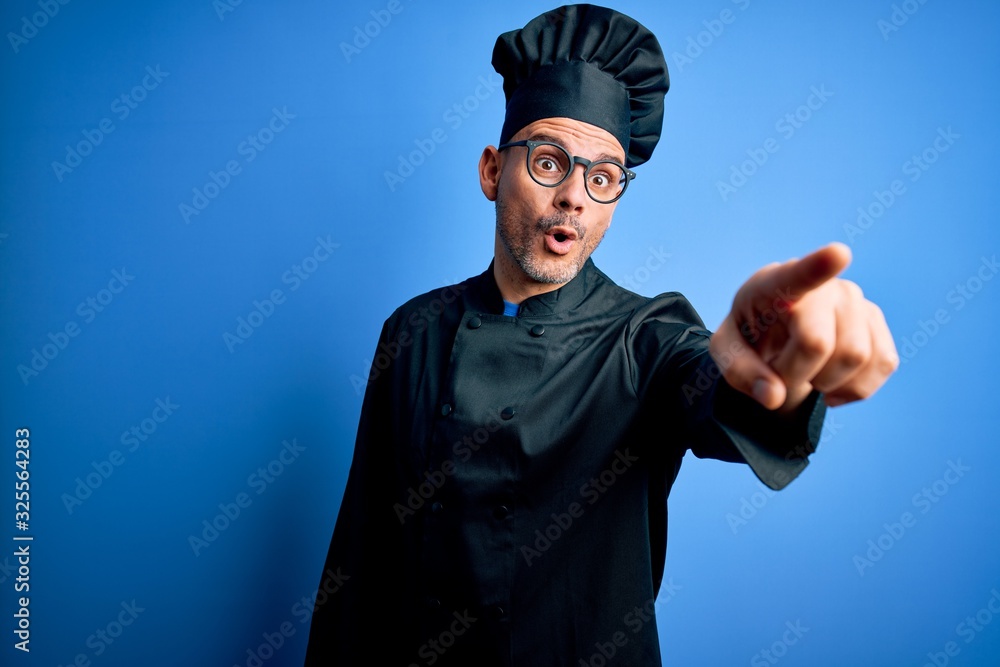 Young handsome chef man wearing cooker uniform and hat over isolated blue background Pointing with finger surprised ahead, open mouth amazed expression, something on the front Young handsome chef man wearing cooker uniform and hat over isolated blue background Pointing with finger surprised ahead, open mouth amazed expression, something on the front