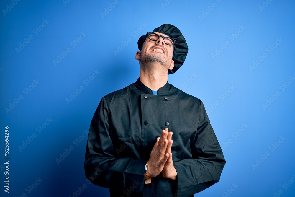 Young handsome chef man wearing cooker uniform and hat over isolated blue background begging and praying with hands together with hope expression on face very emotional and worried. Begging. Young handsome chef man wearing cooker uniform and hat over isolated blue background begging and praying with hands together with hope expression on face very emotional and worried. Begging.
