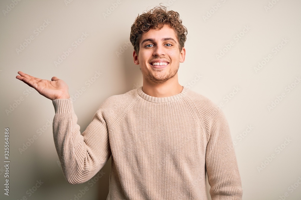 Young blond handsome man with curly hair wearing casual sweater over white background smiling cheerful presenting and pointing with palm of hand looking at the camera. Young blond handsome man with curly hair wearing casual sweater over white background smiling cheerful presenting and pointing with palm of hand looking at the camera.