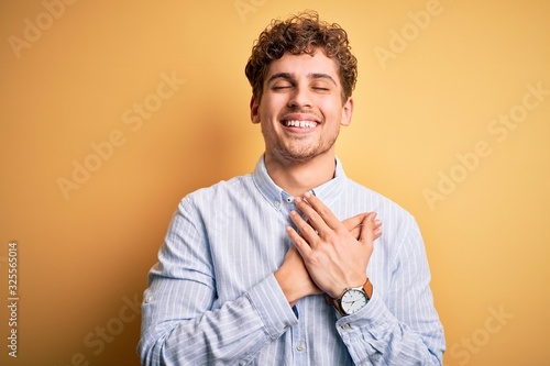 Young blond handsome man with curly hair wearing striped shirt over yellow background smiling with hands on chest with closed eyes and grateful gesture on face. Health concept.