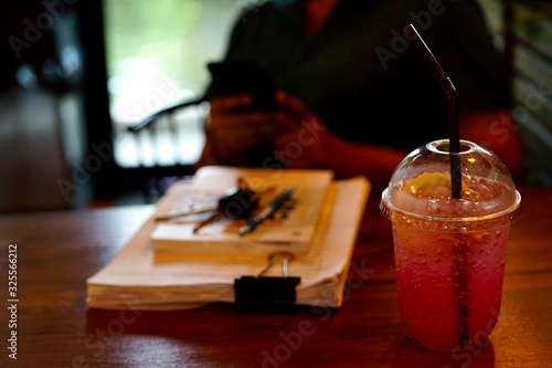 Red sweetened water mixed with soda and lemon is cool drink in a clear plastic glass placed next to the document.The front of the woman who is texting on a mobile phone.