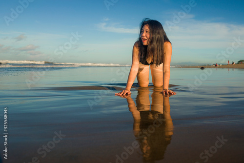natural lifestyle portrait of young attractive and happy Asian Japanese woman in swimsuit kneeling on sand at beautiful beach paradise feeling relaxed and cheerful enjoying holidays
