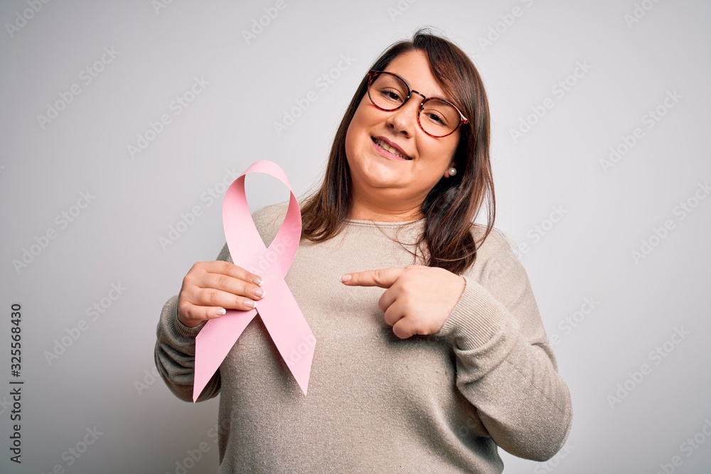 Beautiful plus size woman holding breast cancer awareness pink ribbon over isolated background very happy pointing with hand and finger Beautiful plus size woman holding breast cancer awareness pink ribbon over isolated background very happy pointing with hand and finger