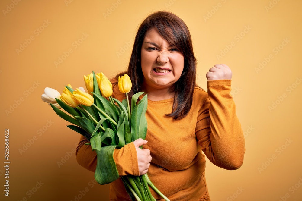 Beautiful plus size woman holding romantic bouquet of natural tulips flowers over yellow background angry and mad raising fist frustrated and furious while shouting with anger. Rage and aggressive Beautiful plus size woman holding romantic bouquet of natural tulips flowers over yellow background angry and mad raising fist frustrated and furious while shouting with anger. Rage and aggressive