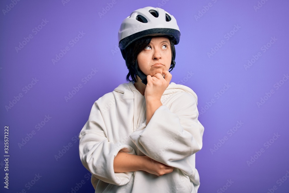 Young down syndrome cyclist woman wearing security bike helmet over purple background with hand on chin thinking about question, pensive expression. Smiling with thoughtful face. Doubt concept. Young down syndrome cyclist woman wearing security bike helmet over purple background with hand on chin thinking about question, pensive expression. Smiling with thoughtful face. Doubt concept.