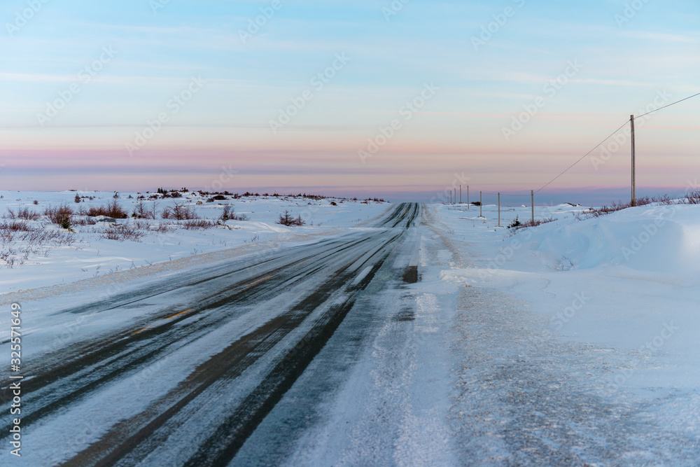A two-lane country road that is paved with snow drifting across it. There's snow on both sides of the road in the barren land area. Your eyes extend to the end of the road with a pink evening sky.
