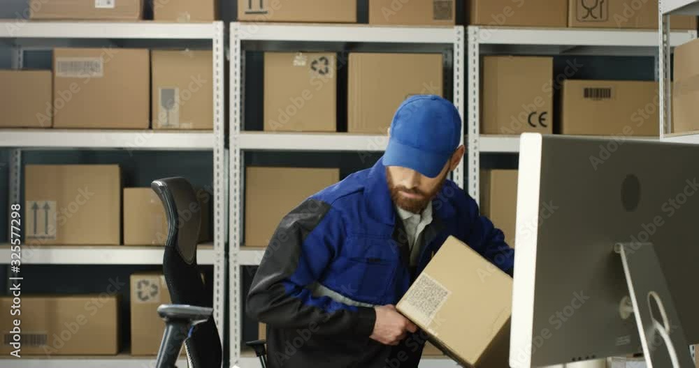 Caucasian man courier taking carton parcel from shelf and bringing to table. Postman registering and scanning box with scanner at post office. Mailman filling invoice and working at computer. Caucasian man courier taking carton parcel from shelf and bringing to table. Postman registering and scanning box with scanner at post office. Mailman filling invoice and working at computer.