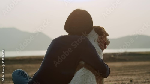 Back view of Asian girl sitting hug retriever dog and stroking with love on the beach at sunset