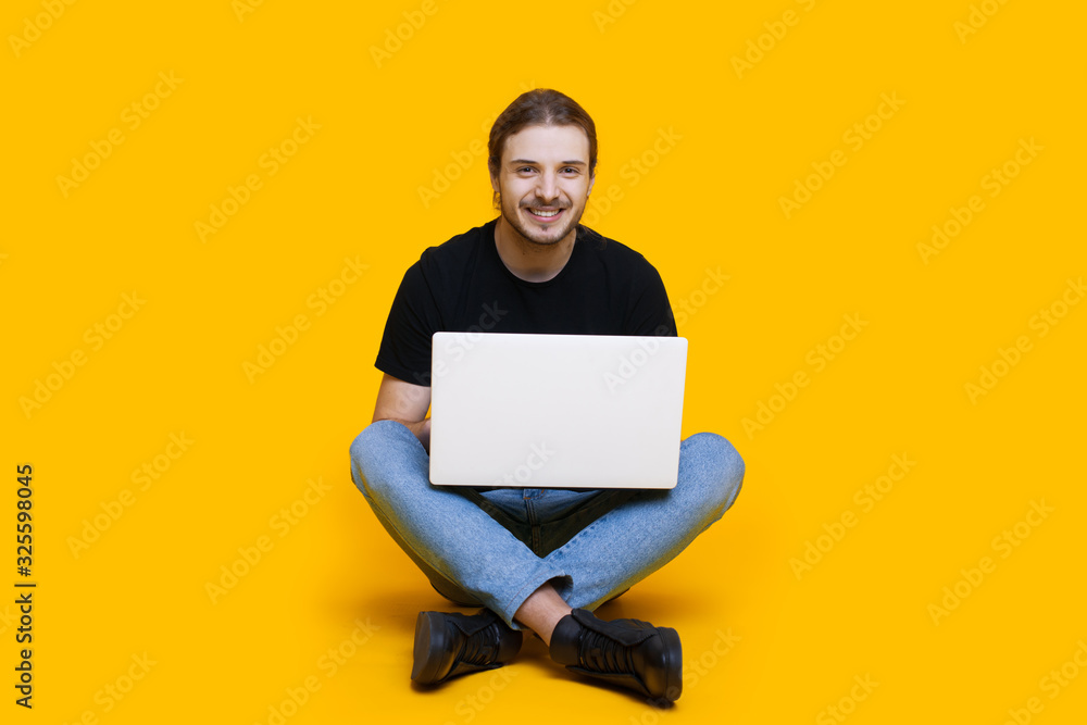 Caucasian unshaven man with long hair sitting with crossed legs typing something at the computer while posing on a yellow background Caucasian unshaven man with long hair sitting with crossed legs typing something at the computer while posing on a yellow background