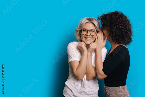 Curly haired caucasian girl is talking something secret to her blonde friend who is wearing eyeglasses and posing on a blue blank space background