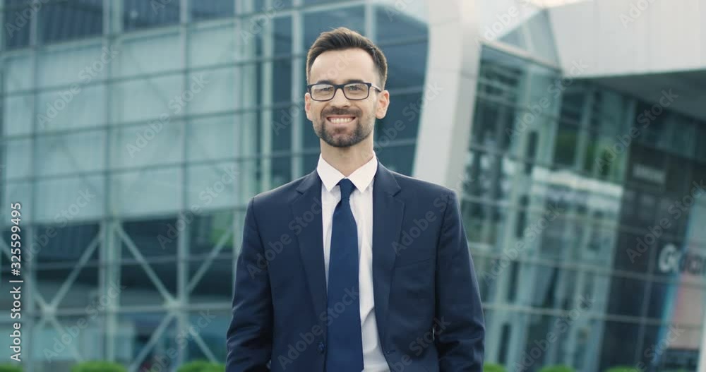 Portrait of young Caucasian handsome man in suit, tie and glasses smiling to camera and standing outdoor at big glass urban building of aeroport or business center. Successful businessman concept. Portrait of young Caucasian handsome man in suit, tie and glasses smiling to camera and standing outdoor at big glass urban building of aeroport or business center. Successful businessman concept.