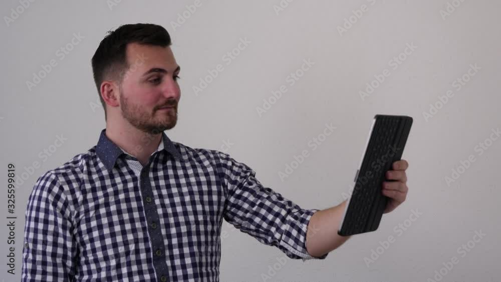 Young man in a shirt in front of a white wall searches for a problem online with his tablet and finds it Young man in a shirt in front of a white wall searches for a problem online with his tablet and finds it