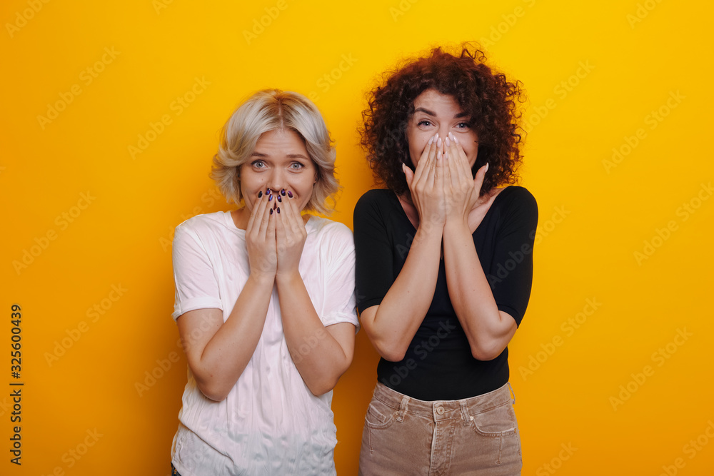 Caucasian curly haired brunette is feeling surprised near her blonde sister covering their mouths with hands on a yellow background Caucasian curly haired brunette is feeling surprised near her blonde sister covering their mouths with hands on a yellow background