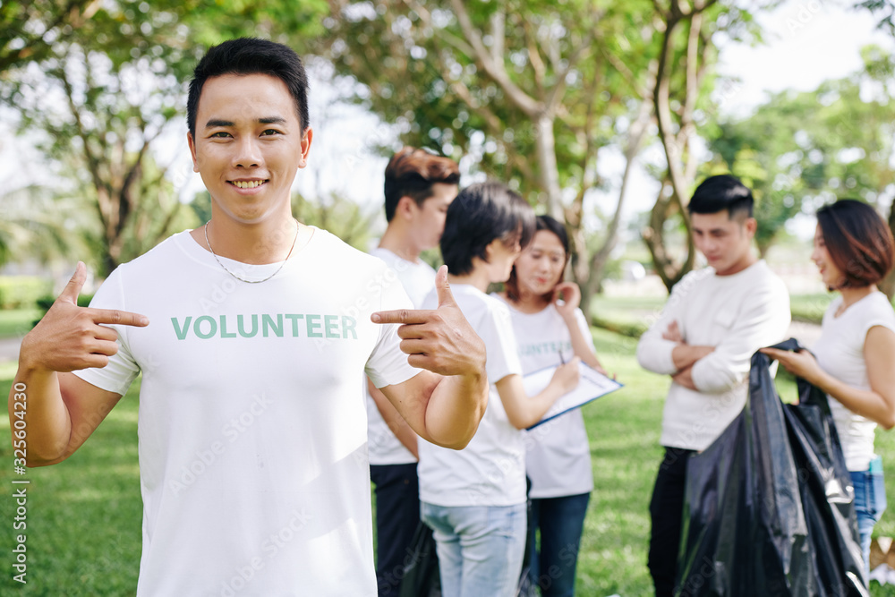 Smiling young Vietnamese man pointing at volunteer inscription on his t-shirt while his team distributing work in background Smiling young Vietnamese man pointing at volunteer inscription on his t-shirt while his team distributing work in background