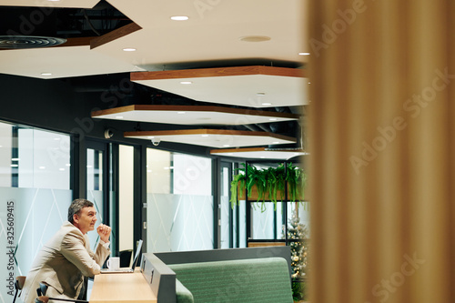 Tableau sur toile Smiling mature businessman drinking mug of coffee and working on laptop in moder