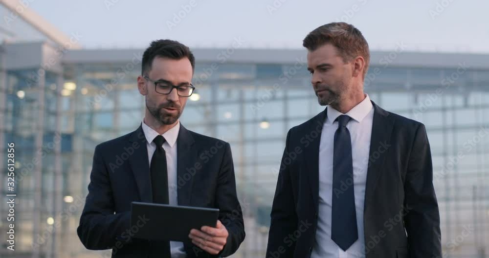 Caucasian two handsome stylish businessmen walking together outdoor and talking. Man demonstrating something on tablet device to male colleague. Business partner showing to co-worker computer screen. Caucasian two handsome stylish businessmen walking together outdoor and talking. Man demonstrating something on tablet device to male colleague. Business partner showing to co-worker computer screen.
