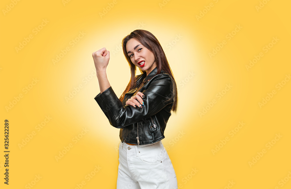 Feminism, girl power, and the concept of equal rights and independence for women. Caucasian young woman in a leather jacket shows biceps. Isolate. Yellow background. Copy Feminism, girl power, and the concept of equal rights and independence for women. Caucasian young woman in a leather jacket shows biceps. Isolate. Yellow background. Copy