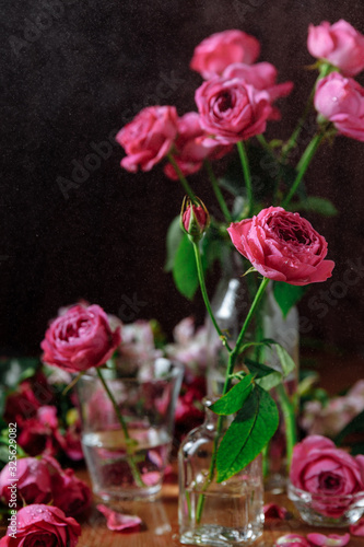 Bunch of pink roses flowers with green leaves and stems in glass vase. Wooden table and dark black background. Still life