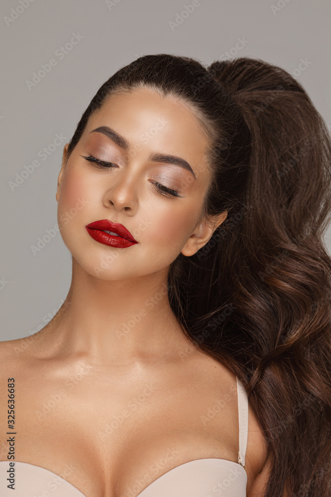 Stock photo portrait of a beautiful young girl with perfect make up and red lips. She has gorgeous thick hair done up in tail. Looking down, wearing bra. Studio shot. Stock photo portrait of a beautiful young girl with perfect make up and red lips. She has gorgeous thick hair done up in tail. Looking down, wearing bra. Studio shot.