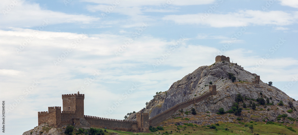 An ancient fortress on top of a mountain against a cloudy sky. Genoese fortress, Sudak, Crimea. Historic old building. Monument of architecture. Panoramic banner.