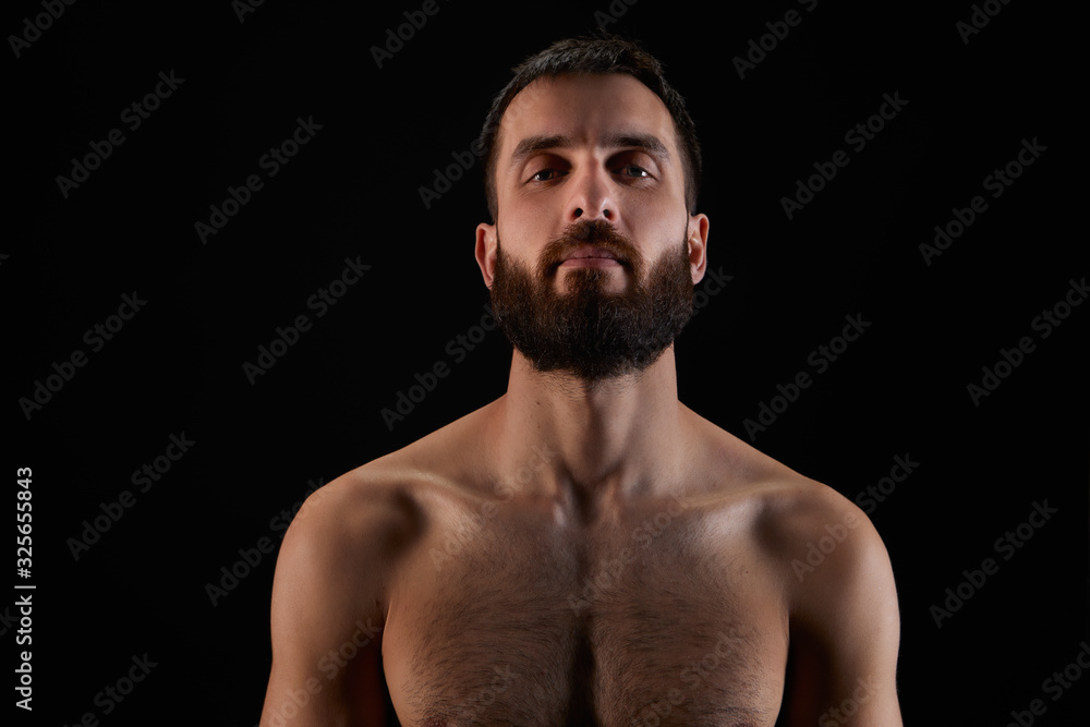 Fototapeta premium Caucasian young man with beard, smiling subtly, shirtless, muscular body, on black background looking straight ahead, horizontal