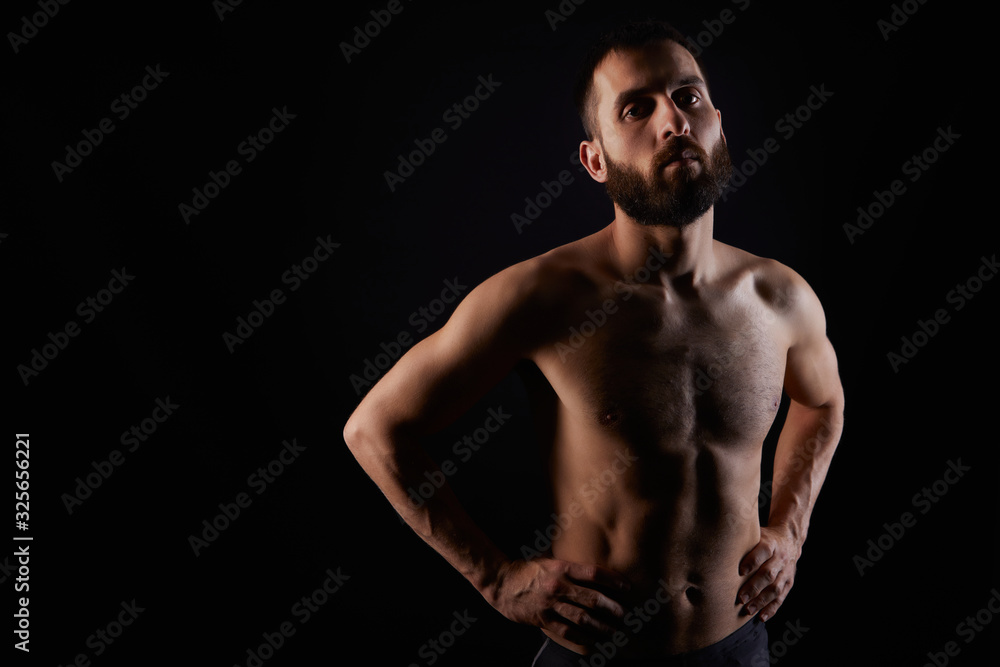 Caucasian young man with beard, serious, shirtless, muscular body, on black background looking straight ahead with hands on waist, horizontal