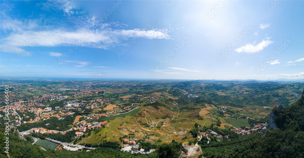 Fototapeta premium High top aerial view Italy landscape in summer, hills and farms, fields and forests, in the far mountains. panoramic wallpaper. In the distant city of Rimini and the sea. San Marino