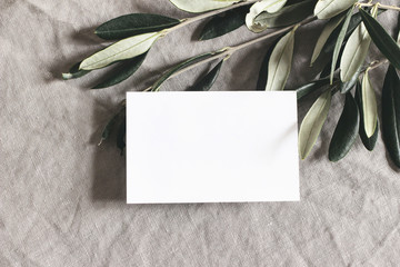 Closeup of blank greeting, business card and green olive leaves, branches on linen tablecloth background. Moody feminine wedding stationery mock-up scene. Flat lay, top view. Mediterranean design.