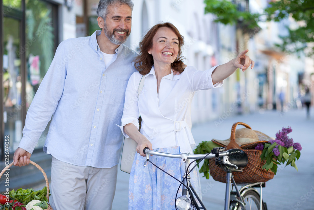 Happy mid adult married couple returning from groceries shopping on a farmers market, walking through town. Woman is pushing a bicycle and pointing into distance