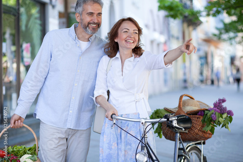 Obraz na płótnie Happy mid adult married couple returning from groceries shopping on a farmers market, walking through town. Woman is pushing a bicycle and pointing into distance w salonie