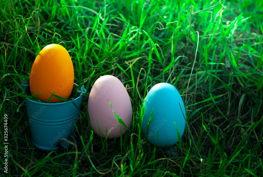 an orange Easter egg in a small bucket of blue that stand among the grass and next to it are two pink and blue eggs an orange Easter egg in a small bucket of blue that stand among the grass and next to it are two pink and blue eggs