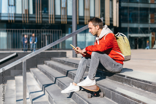 Handsome smiling hipster with wooden longboard sitting on the steps and chatting in the mobile phone. Concept of leisure activity. Outdoor urban and street photo. Healthy lifestyle, fashion look.