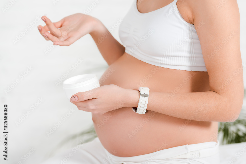 Cropped view of pregnant girl holding jar with cosmetic cream while sitting on bed