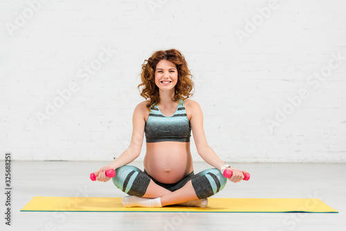 Pregnant woman with dumbbells smiling at camera while sitting on fitness mat at home
