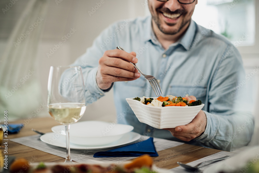 Close-up of happy man taking salad from a bowl while having lunch at dining table.