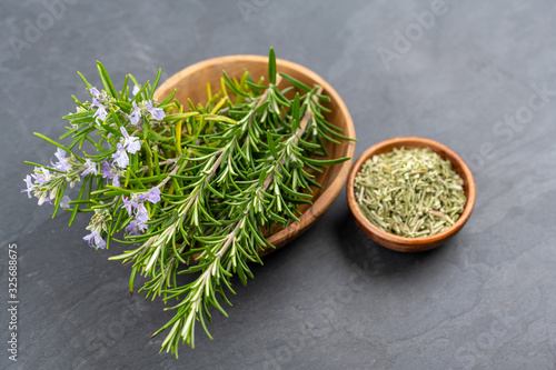 Purple blooming rosemary and fresh rosemary twigs in an olive wood bowl and a small bowl of whole dried rosemary on a black slate background