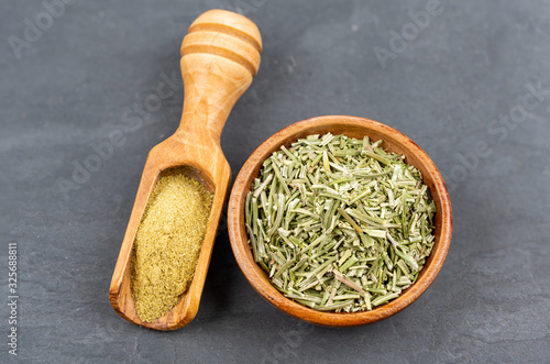 A small bowl with whole dried rosemary and a spice shovel with ground rosemary on a black slate background