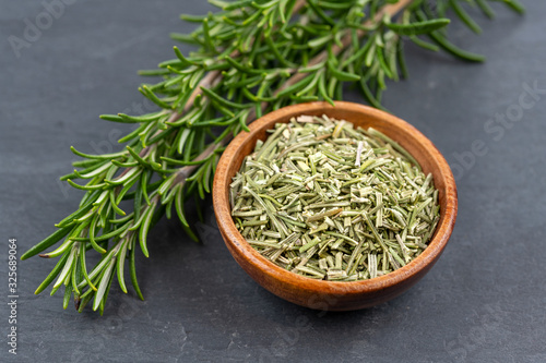Fresh rosemary twigs and a wooden bowl with dried rosemary on a black slate background