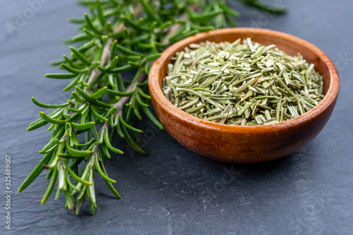 Fresh rosemary twigs and a wooden bowl with dried rosemary on a black slate background
