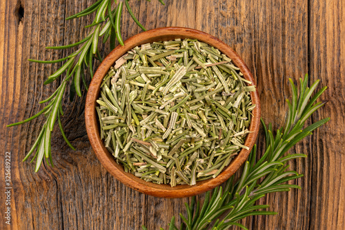 A wooden bowl filled with whole dried rosmary and two rosemary twigs on a rustic wooden background