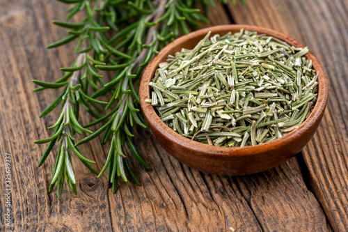 Close-up view of a wooden bowl filled with whole dried rosmary and two rosemary twigs on a rustic wooden background