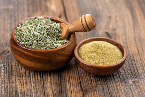 A wooden bowl filled with whole dried rosemary with a spice shovel and a small wooden bowl filled with ground rosemary on a rustic wooden background