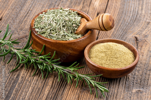 A wooden bowl with whole dried rosemary with a spice shovel, a fresh rosemary twig and a small wooden bowl filled with ground rosemary on a rustic wooden background