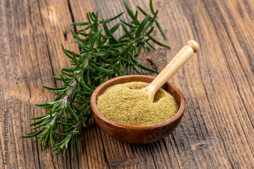 A small wooden bowl with ground rosemary and a small spice shovel and fresh rosemary twigs on a rustic wooden background