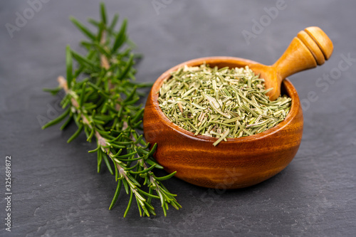 A wooden bowl with whole dried rosemary and a spice shovel and fresh rosemary twigs on a black slate background