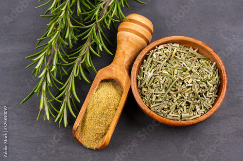 Fresh rosemary twigs, a spice shovel with ground rosemary and a wooden bowl with dried rosemary on a black slate background