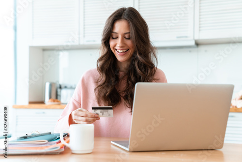 Woman sit indoors at home using laptop holding credit card.